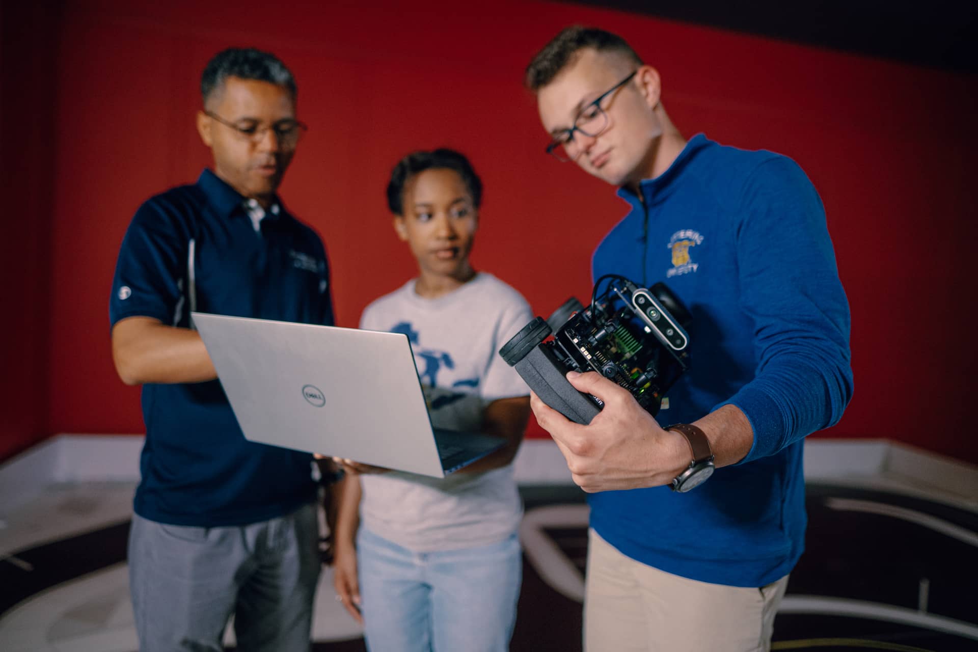 A Kettering professor points at a laptop that a student holds. Another student holds and scrutinizes a self-driving robot.