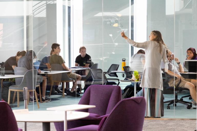 Students engage in a collaborative group discussion guided by an instructor in a glass-walled study room at the Learning Commons.