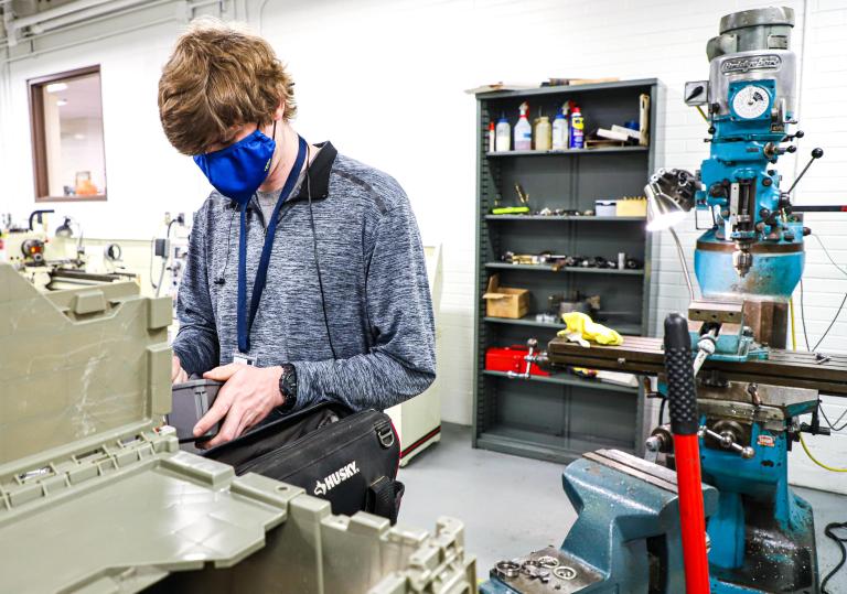 A masked Kettering University student works on piece of machinery.