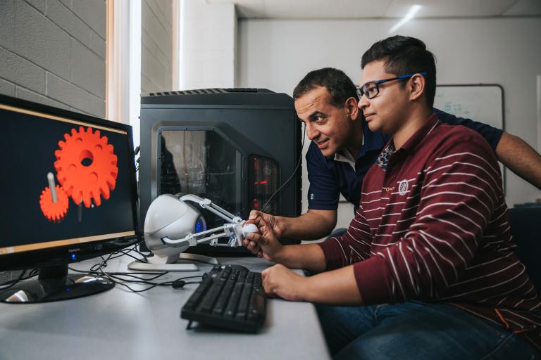 A professor leans over to help a student working at a computer.