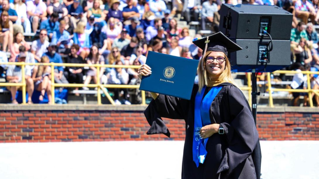 A student in a cap and gown holds up a diploma at Commencement