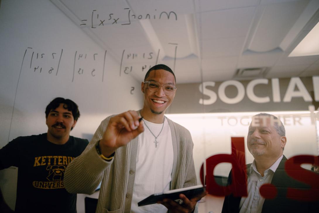 A School of Management Kettering student writes matrix equations on a glass wall. Another student and a professor watch