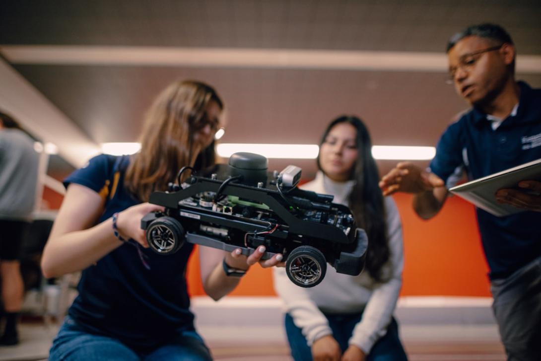 One Kettering student holds a small autonomous vehicle while another watches. A Kettering professor gives instructions.