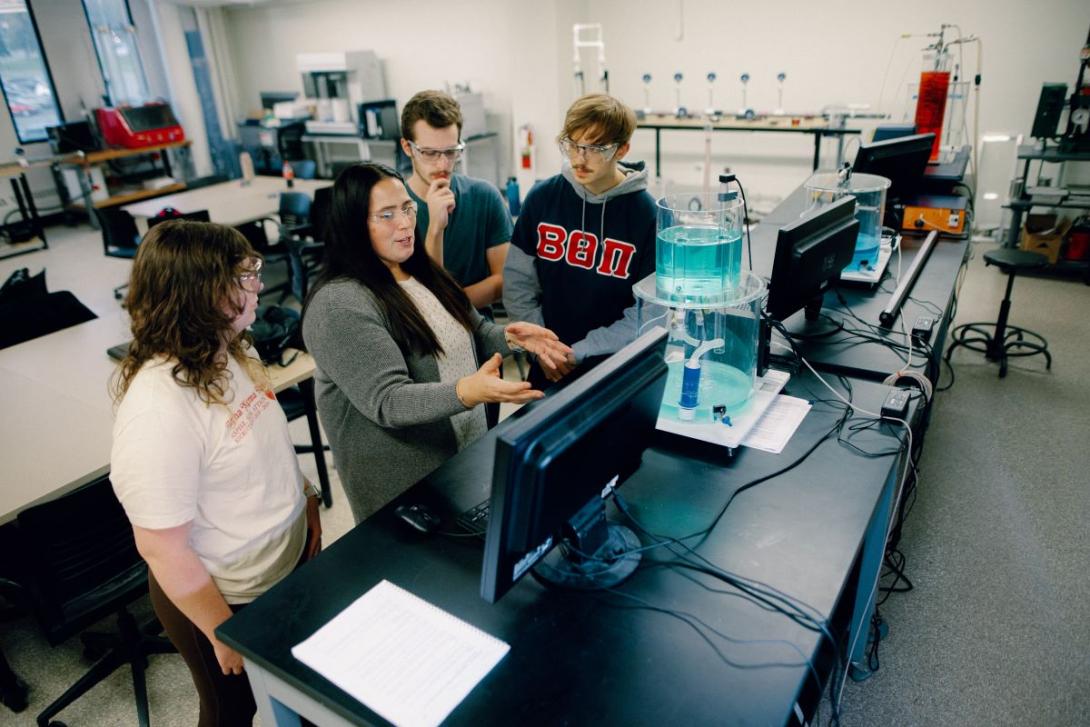 A Kettering professor instructs three students in the Chemical Engineering Lab. On the table is a computer and lab equipment.