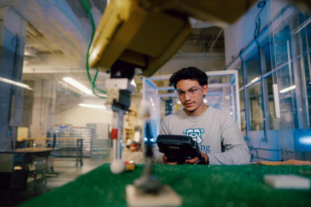 A Kettering student wearing safety goggles uses an industrial robot arm to test materials in the Polymer Lab