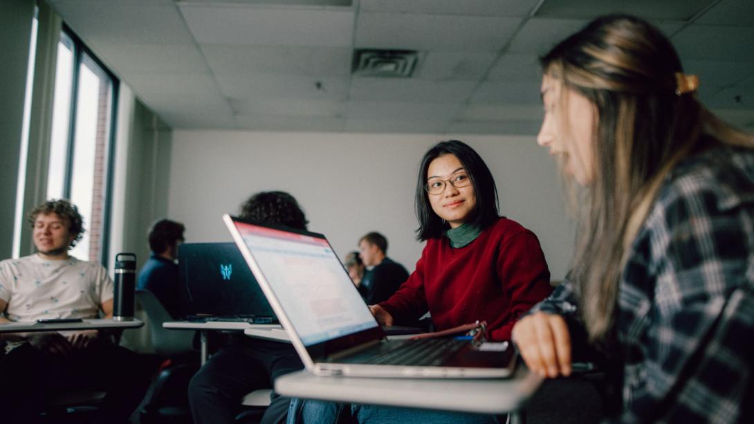 A Kettering student wearing a red sweater looks at another student. They sit in a classroom and each have open laptops