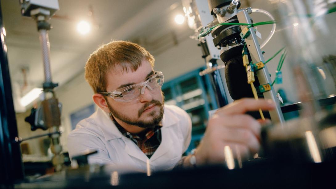 A Kettering chemical engineering student wears safety glasses and a white lab coat and inspects lab equipment