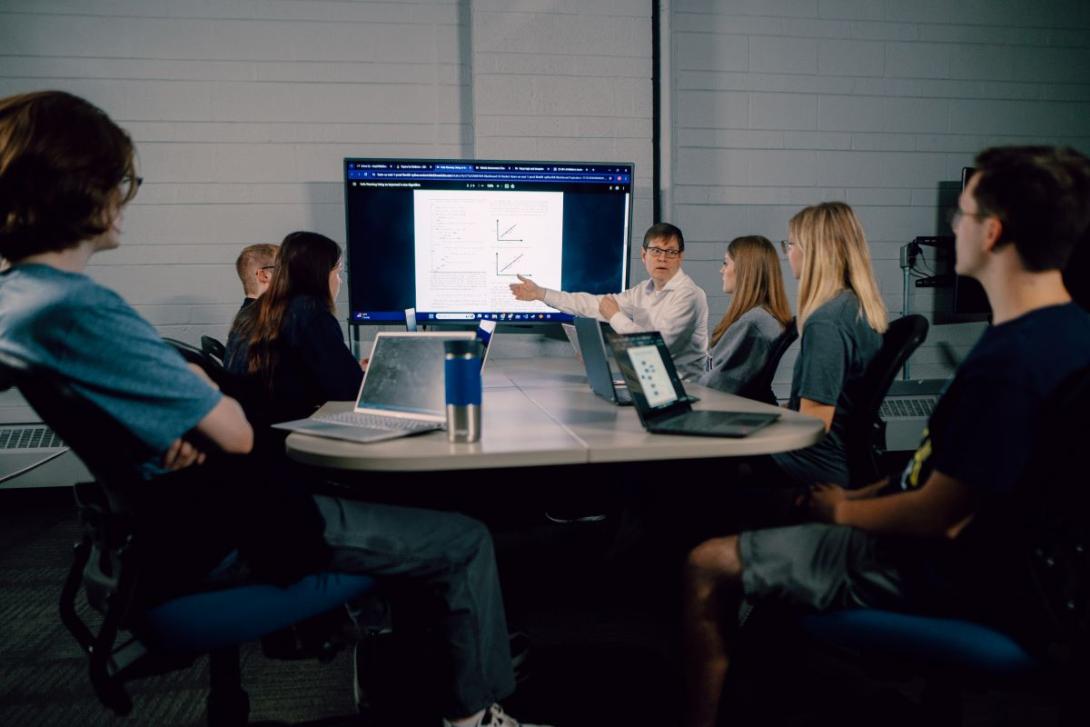 Kettering students watch a professor's computer science presentation. They are seated around a table and have laptops. 