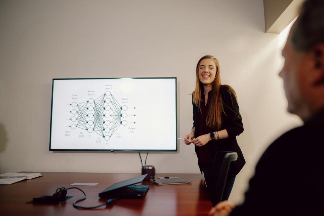 A Kettering School of Management student stands in a conference room. Her presentation is shown via a large monitor.