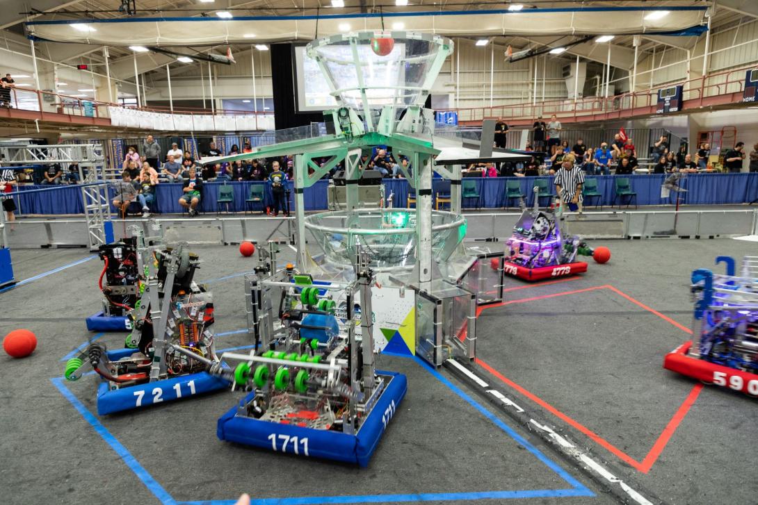 Four FIRST Robotics robots complete tasks in a competition field as an audience watches