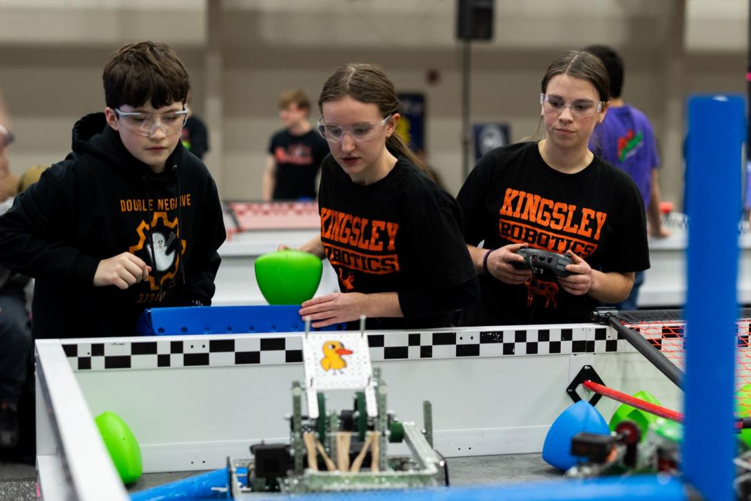 Three high school students wearing safety glasses watch their robot compete at the VEX State Championship at Kettering