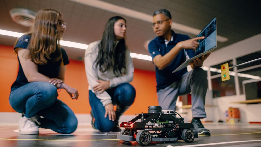 A Kettering professor points to a laptop as two students look on. In front of them is an autonomous electric vehicle