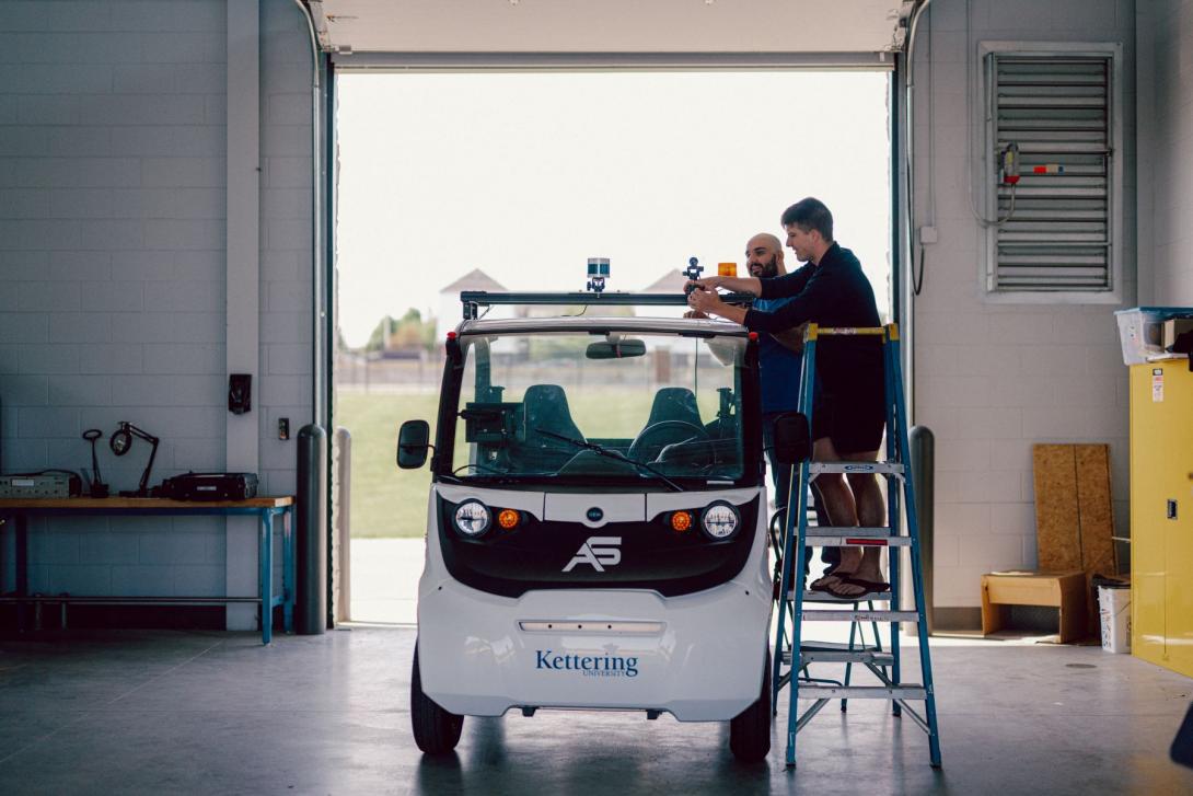 Two Kettering students standing on ladders adjust cameras on top of an electric vehicle in the Mobility Research Center.