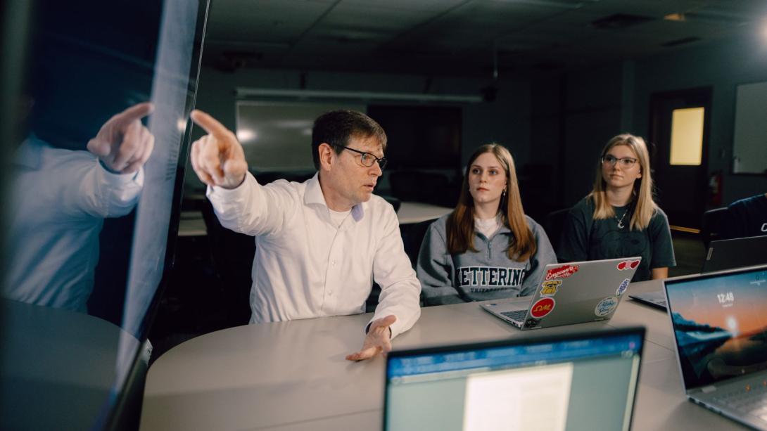 A Kettering professor points at a large television screen while two students look on. Each student has a laptop