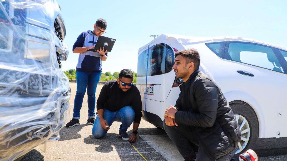 Three Kettering students measure markings on a road at the Mobility Research Center using measuring tape. They stand between two cars.