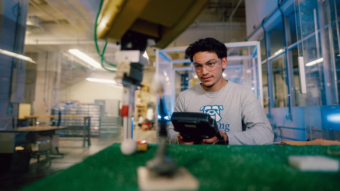 A Kettering industrial engineering student wears safety goggles and controls and industrial robot arm in the Polymer Lab