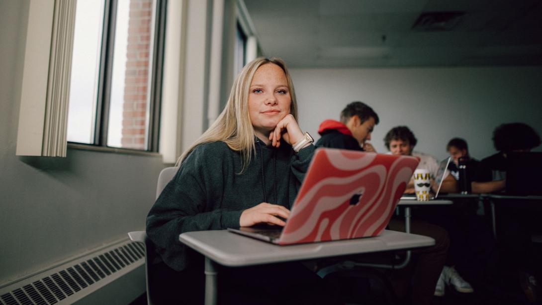 A Kettering management student sits in a classroom. On the desk is a laptop with a pink case.