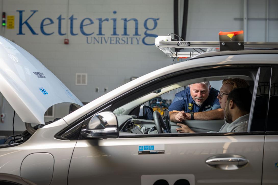 Two Kettering students and a professor look at a car's computer panel in the Mobility Research Center. The car's hood is up