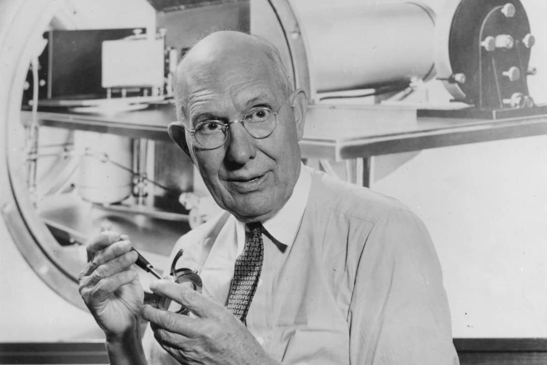 A black-and-white photo of Charles Kettering, sitting in a lab and holding machine parts.