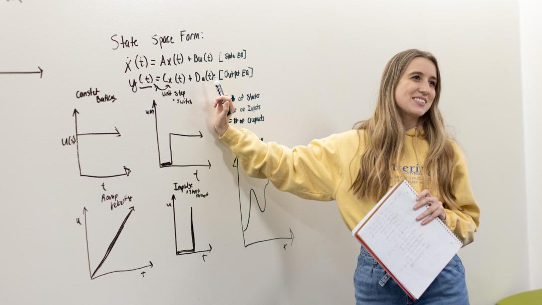 Student standing at a whiteboard with a mathematic problem displayed