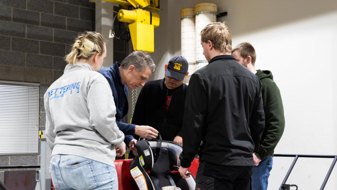 A group of students watch a professor work with a car seat in the Crash Lab