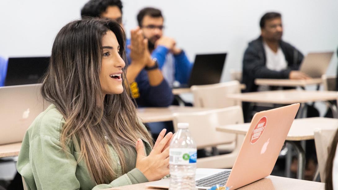 A student sits at a desk with an open laptop in a classroom with other students in background