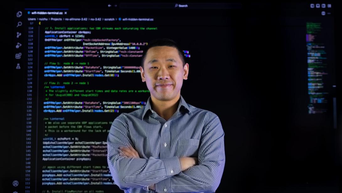 An individual in business attire stands with arms crossed in front of a wall of computer code