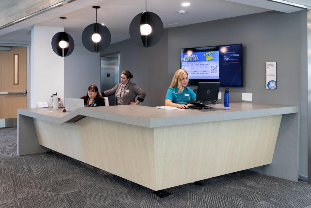 Two Kettering employees and a student worker  work at computers at the Library Services desk in the Learning Commons