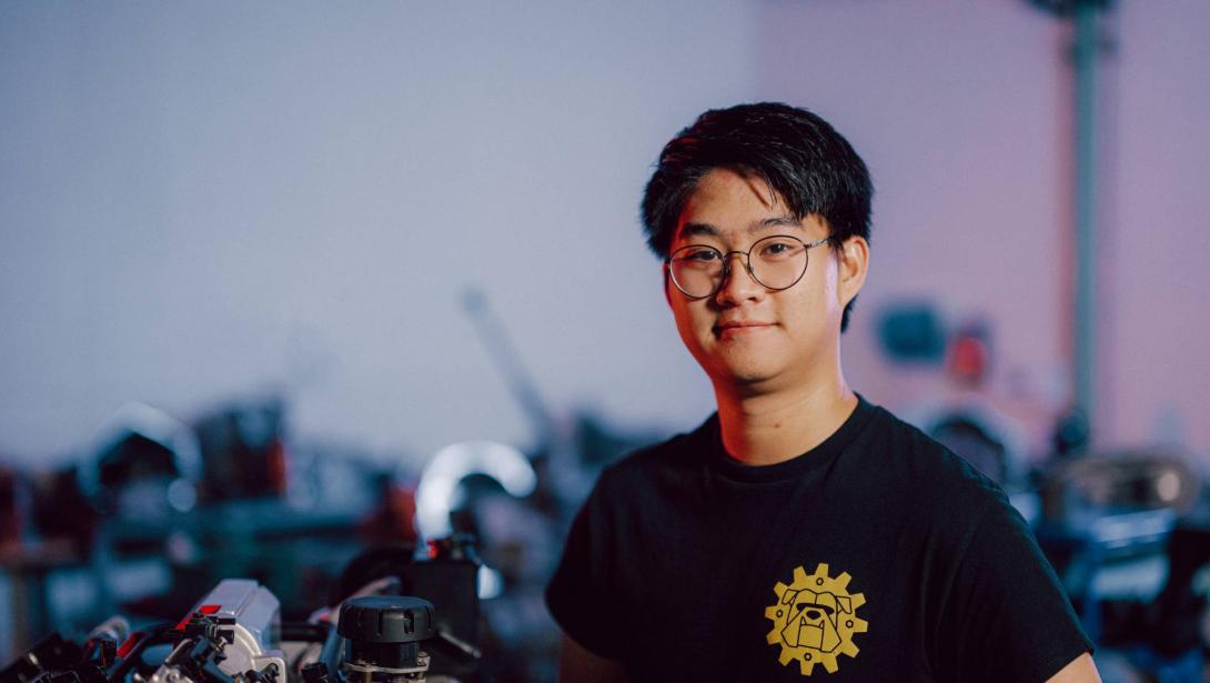 A Kettering student wearing a black shirt stands in the Car Lab alongside an engine component