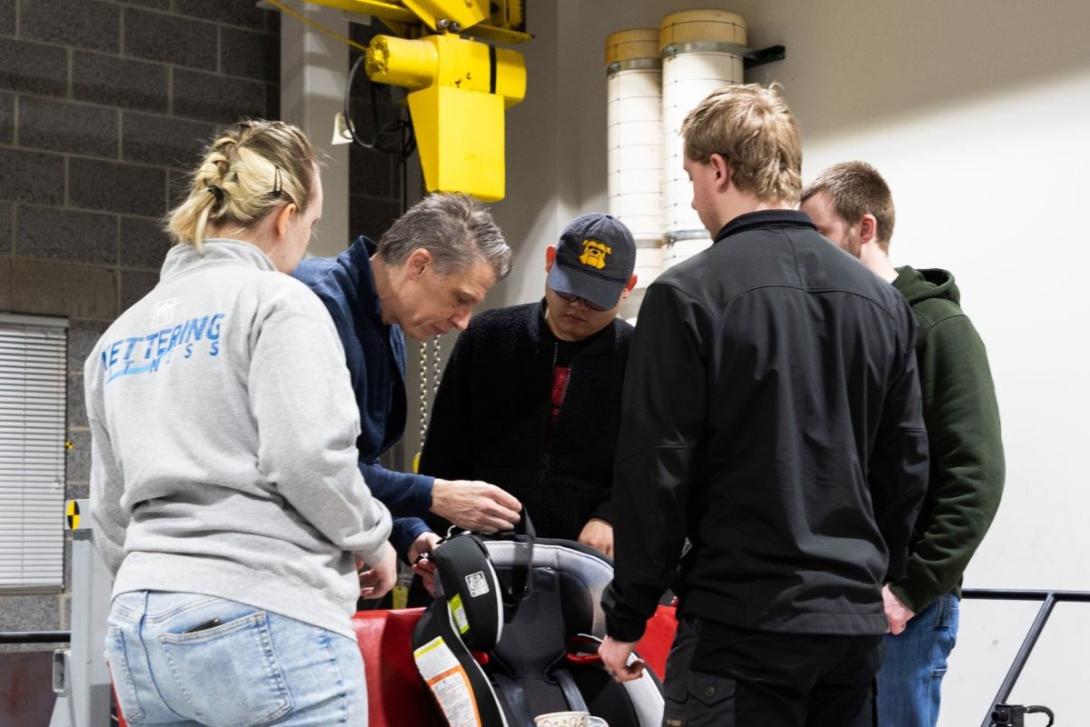 A group of students watch a professor work with a car seat in the Crash Lab