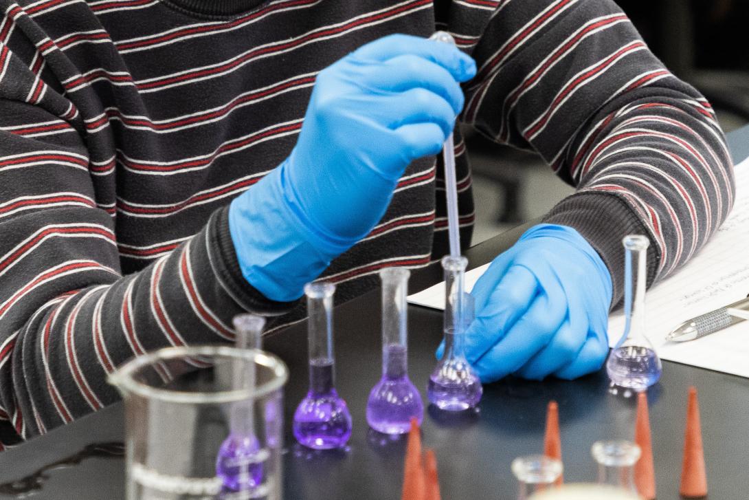 A pair of hands wearing gloves adding liquid to a beaker with a pipette