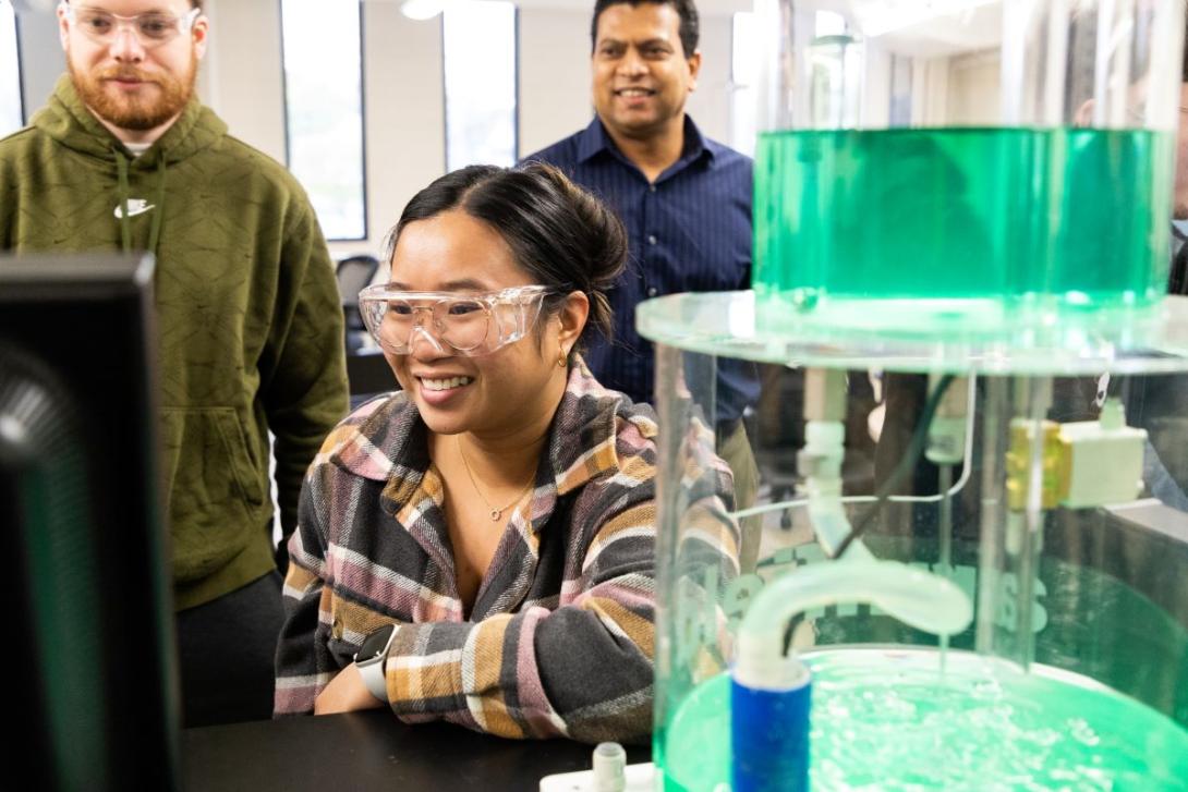 A student wearing googles sits at a computer in a lab with a an experiment in the foreground and two other people standing in the background