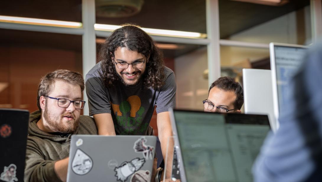 Students gathered around a table looking at and pointing to a laptop