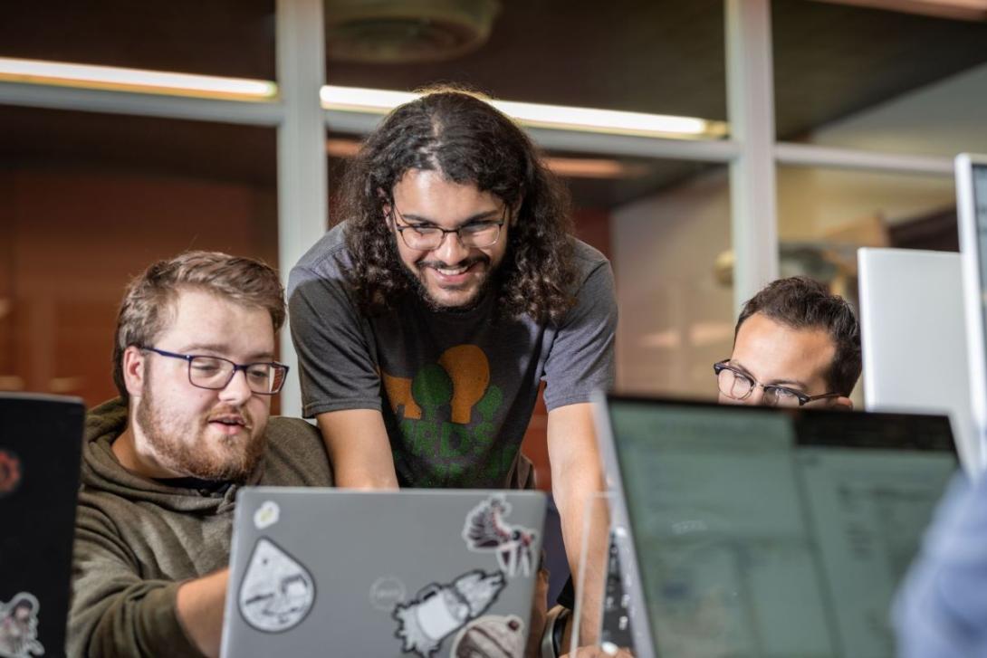 Students gathered around a table looking at and pointing to a laptop