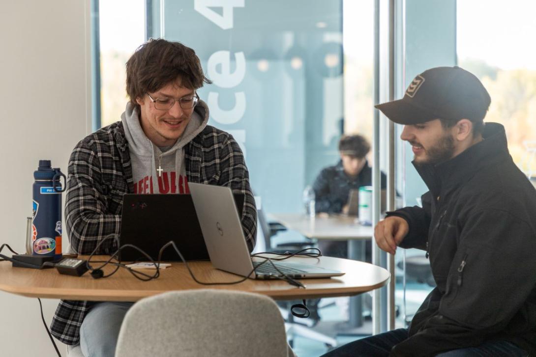 Two students sit at a small table looking at laptops