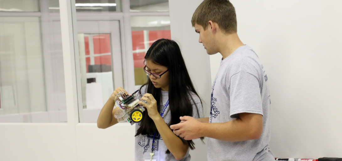 "Two students deeply engaged in hands-on robotics work, carefully examining and adjusting components of a small wheeled robot as part of a collaborative STEM project in a modern classroom setting."