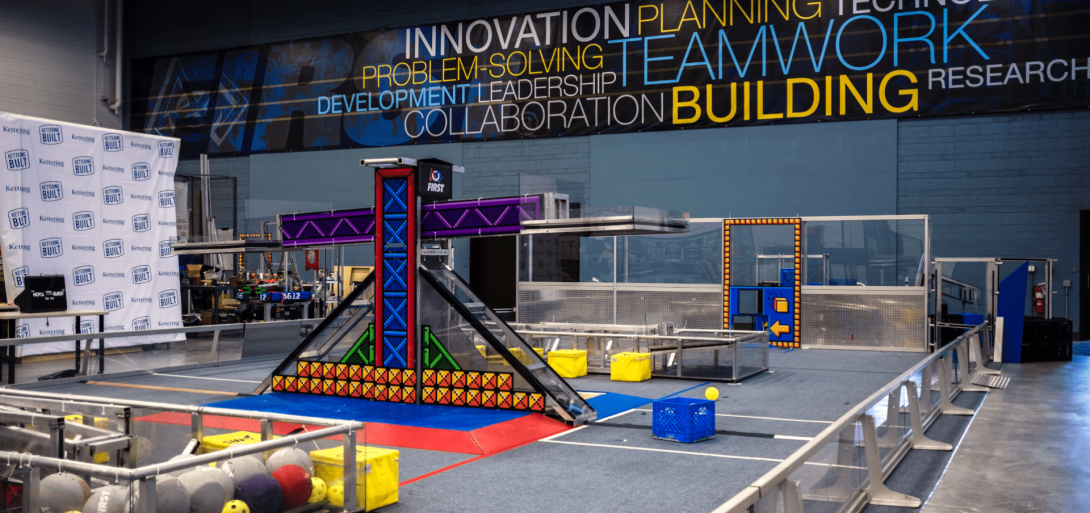 A robotics practice field at Kettering University's FIRST Robotics Community Center, featuring game elements, a central structure, and banners promoting innovation and teamwork.