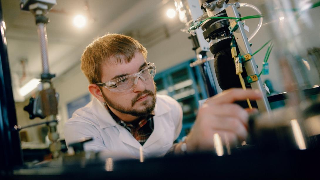 A student in a lab coat and goggles works with equipment