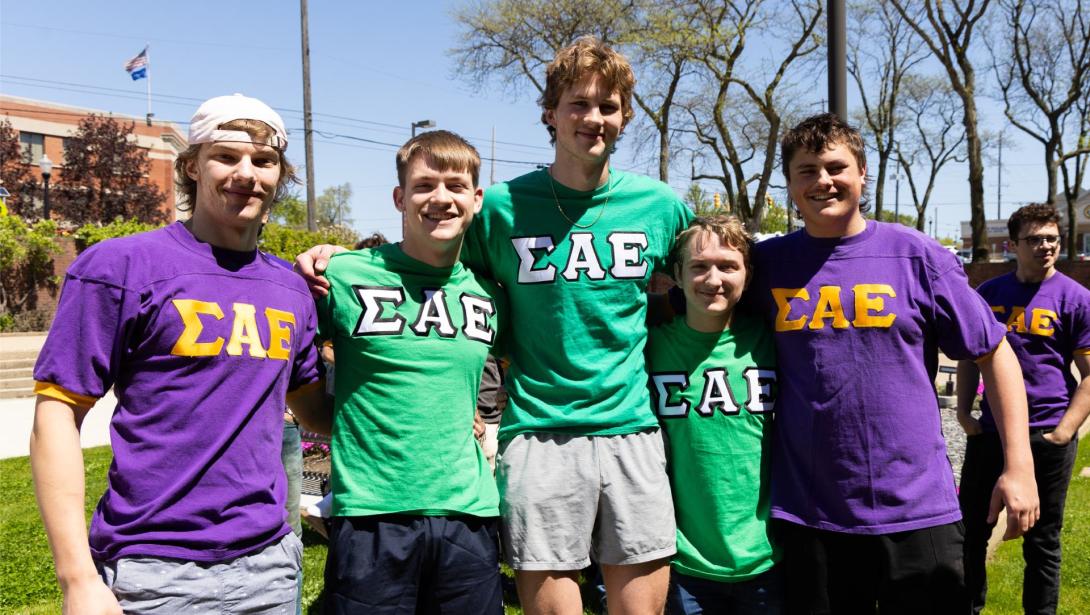 Five Kettering students standing outside smiling while wearing purple and green Sigma Alpha Epsilon fraternity shirts