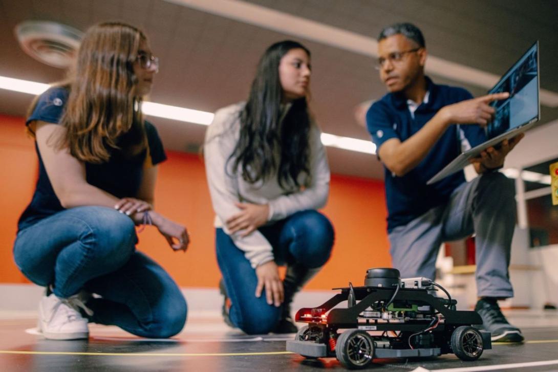 A Kettering professor points to a laptop as two students look on. In front of them is an autonomous electric vehicle