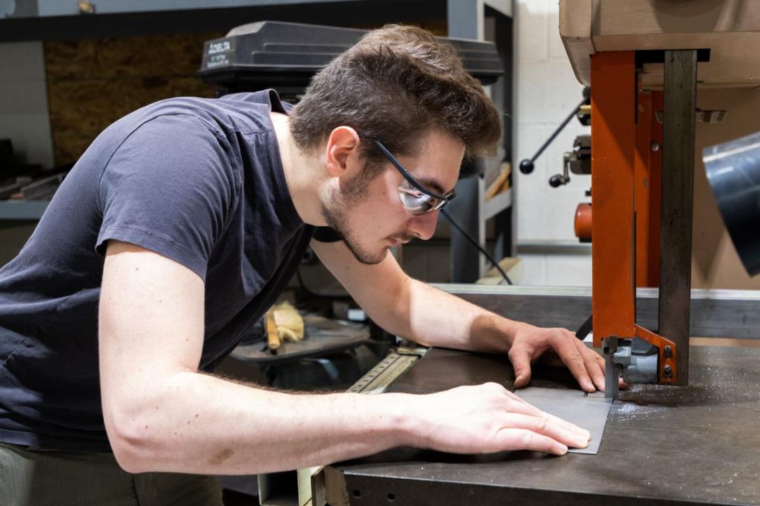 A Kettering student wearing safety glasses cuts a piece of metal with heavy equipment in a lab space