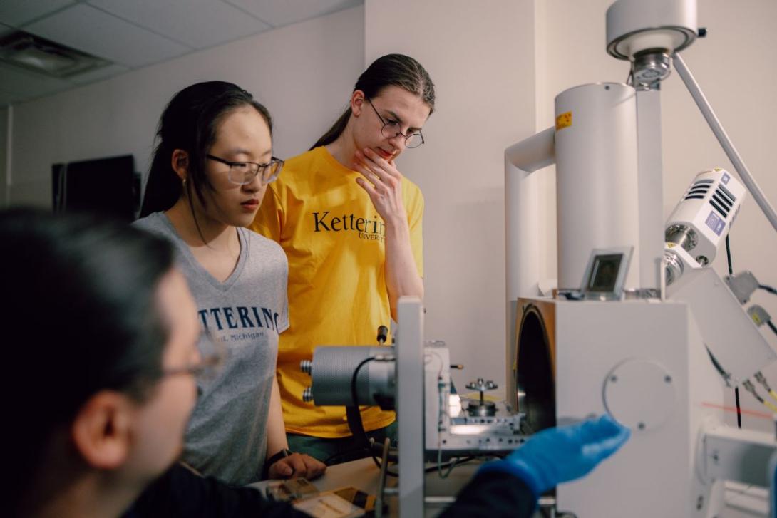 A Kettering professor is seated at an electron microscope instructing two students standing nearby