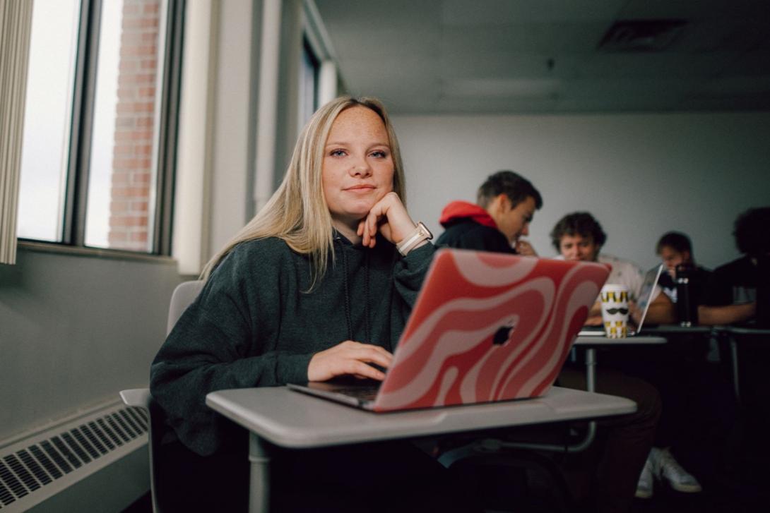 A Kettering management student sits in a classroom. On the desk is a laptop with a pink case.