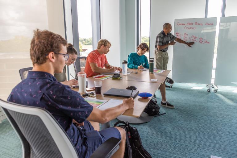 A faculty member guides students in an interactive study session using whiteboards in a dedicated meeting room within the Learning Commons.