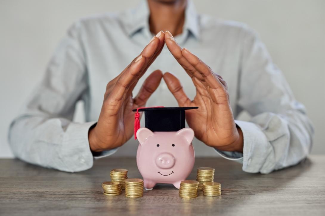 Hands held over a small piggy bank wearing a commencement cap with money stacks around it